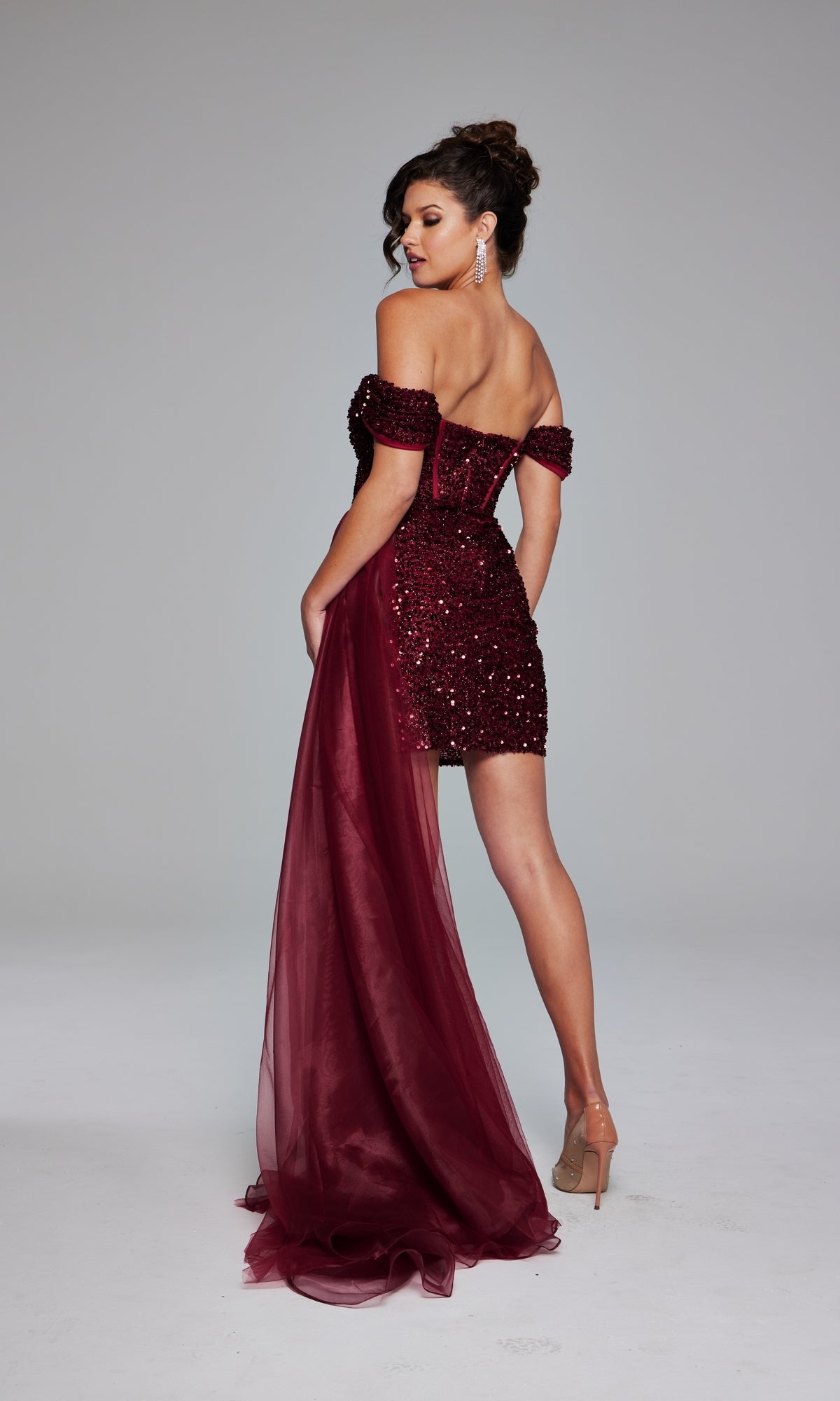 A woman poses with her back to the camera, wearing a burgundy Jovani Off-Shoulder Short Sequin Hoco Dress 39632 with a sheer train.